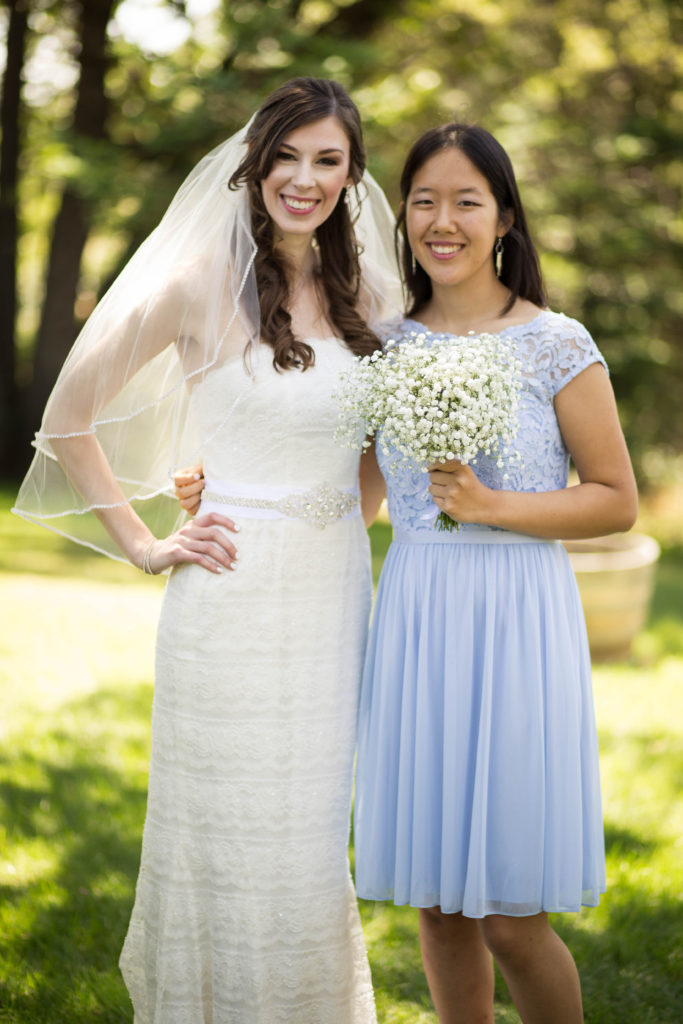 Jenna wearing white wedding dress and veil standing next to bridesmaid Victoria in a blue dress holding a baby breath flower bouquet.