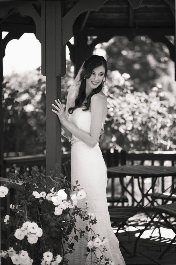 Black and white photo of Jenna in white wedding dress standing beside pillar and a rosebush