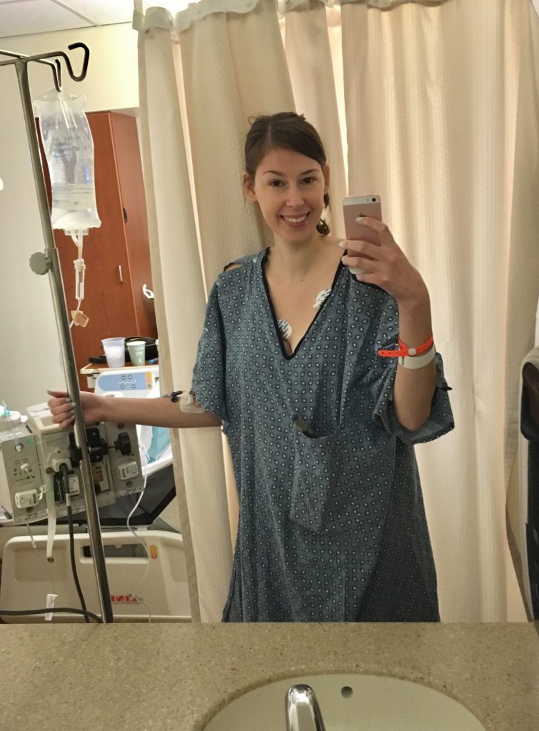 Jenna taking selfie in hospital bathroom wearing blue hospital gown and holding onto IV pole