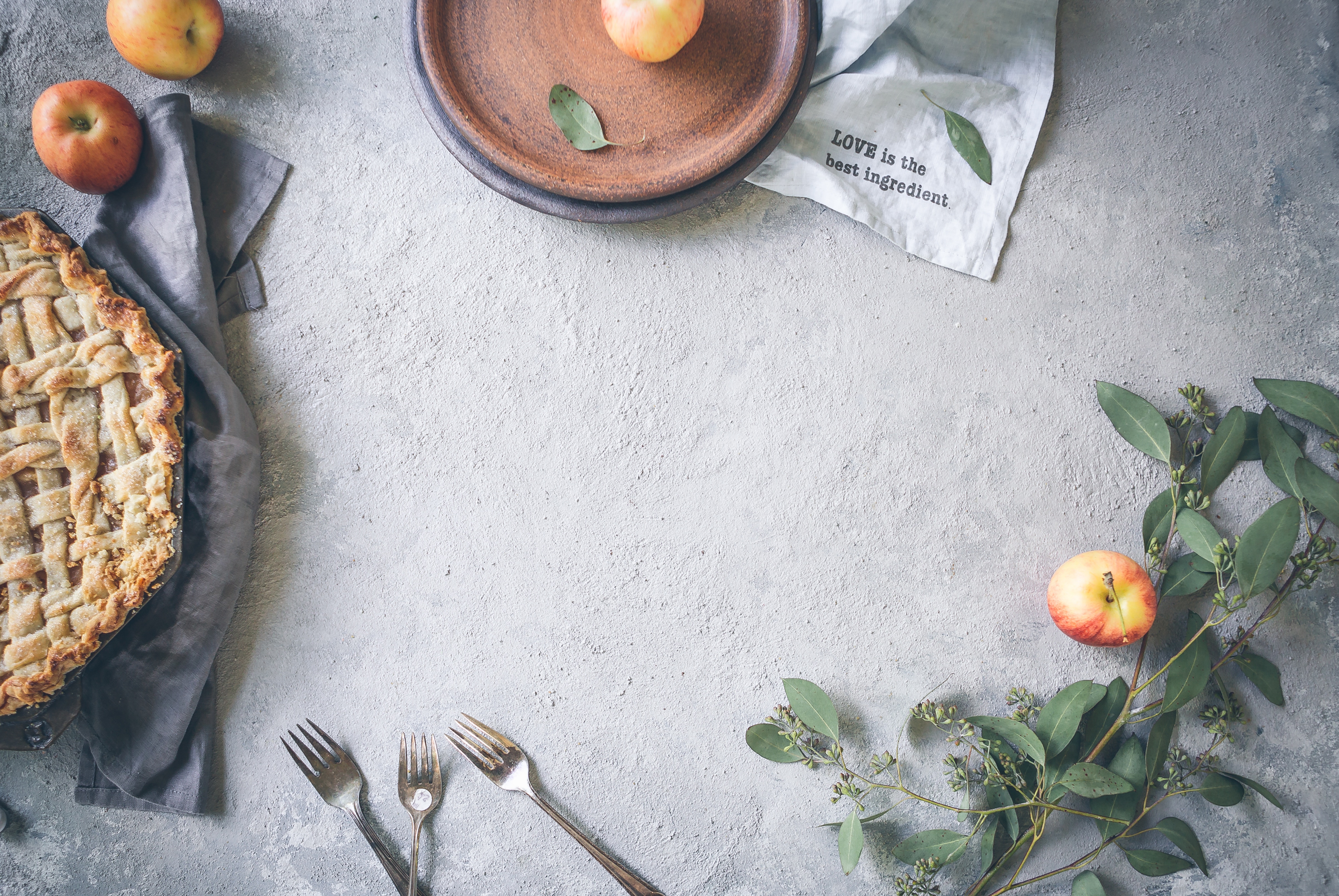 Pie, silverware, wooden bowls, and greenery sitting on grey, cement tabletop