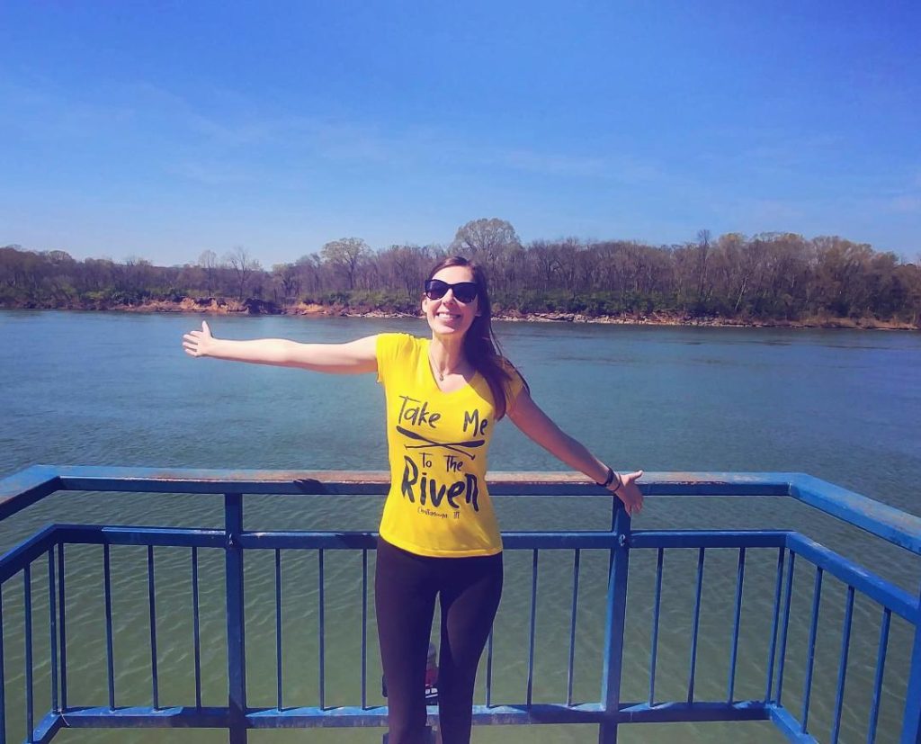 Jenna standing with arms wide in yellow shirt that reads "take me to the river" in front of a blue river