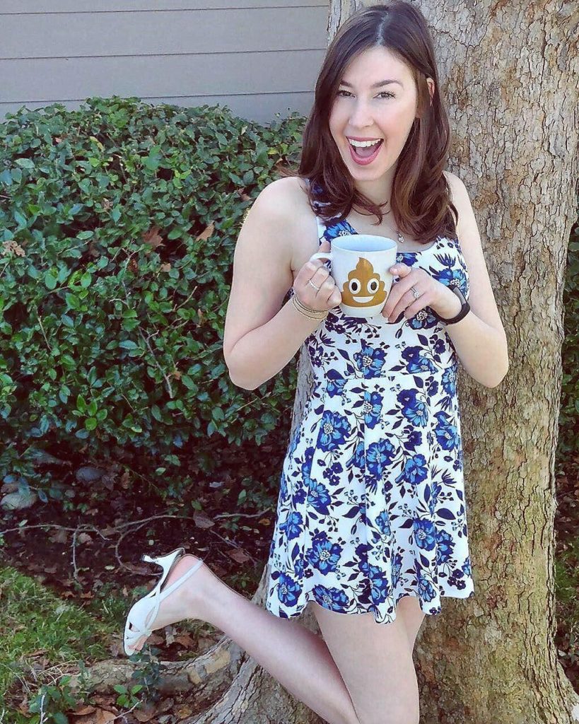 Jenna wearing blue floral dress and white heels holding poop mug beside tree
