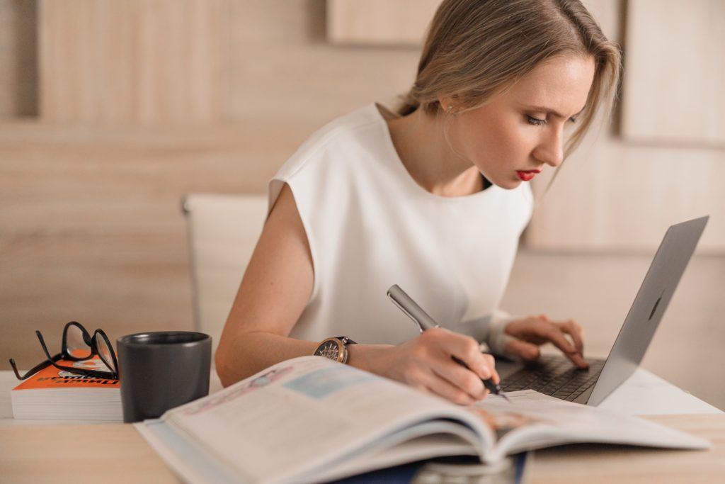 Woman hunched over desk scribbling in a notebook beside a laptop