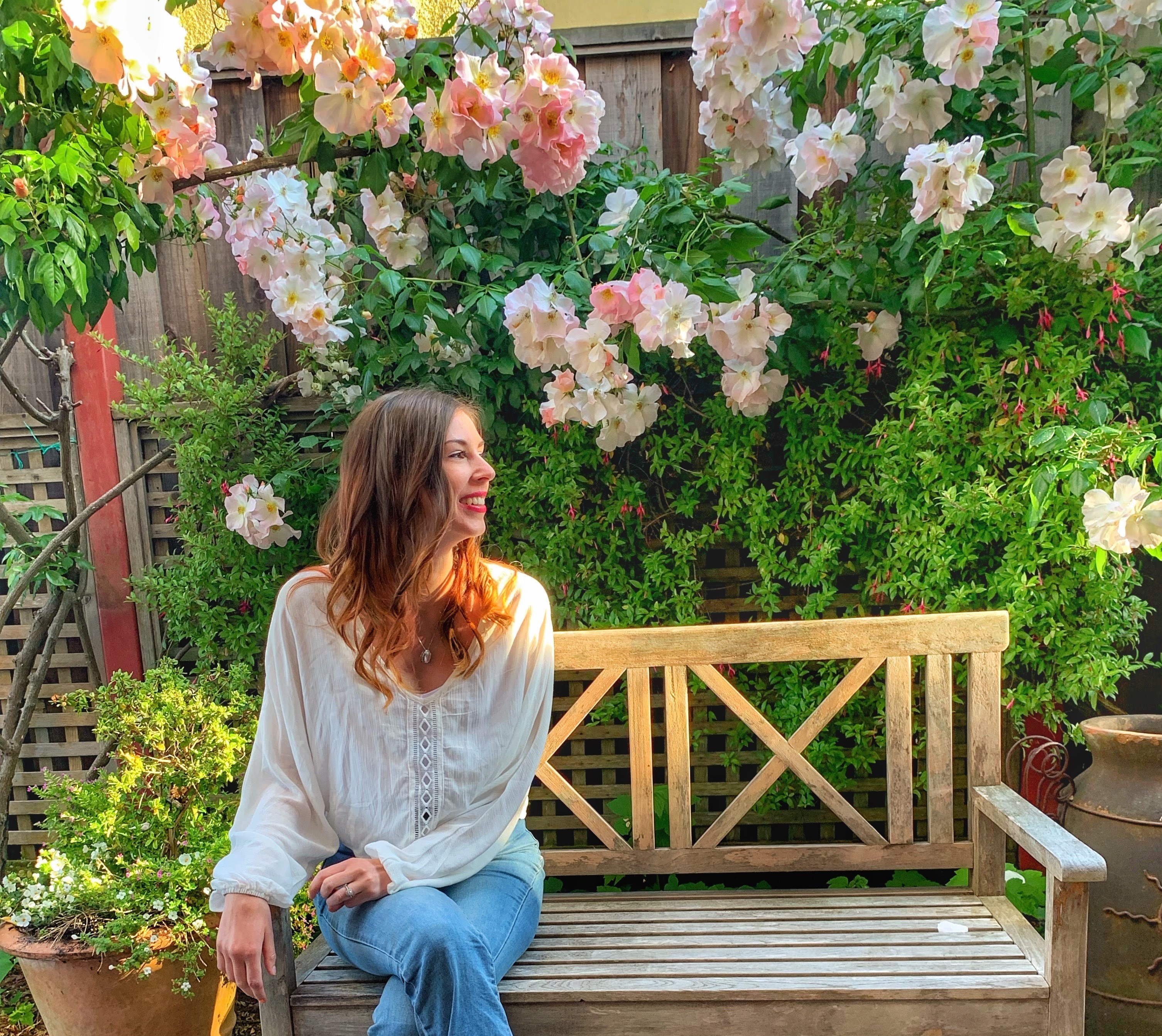Jenna sitting on a bench in front of a wall of flowers smiling toward the sun