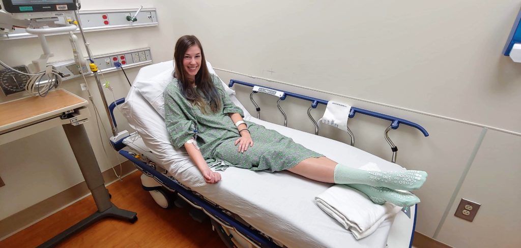 Jenna sitting on hospital bed wearing green gown and IV in arm