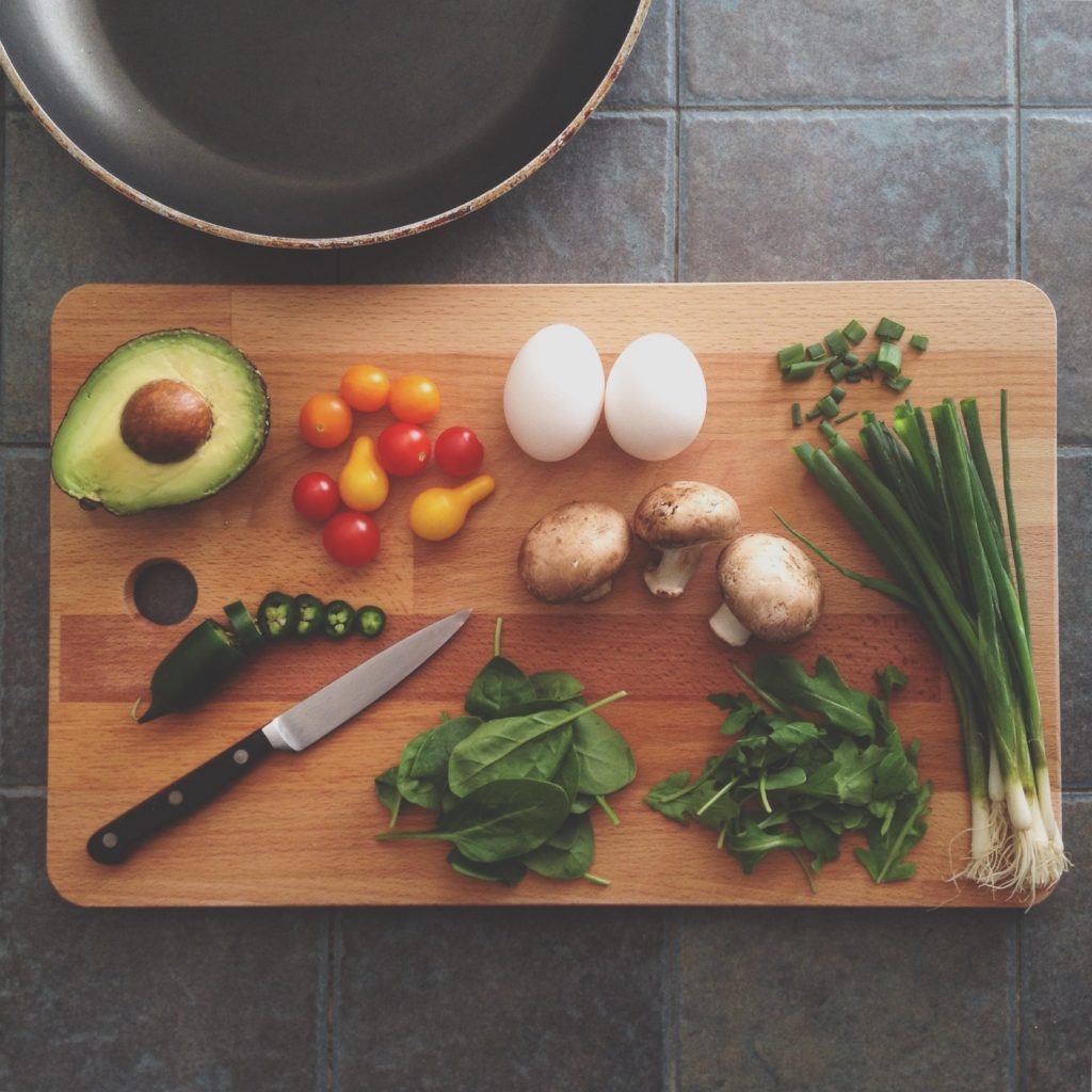 Avocado, eggs, tomatoes, spinach, and other vegetables laying beside knife on a wooden cutting board.